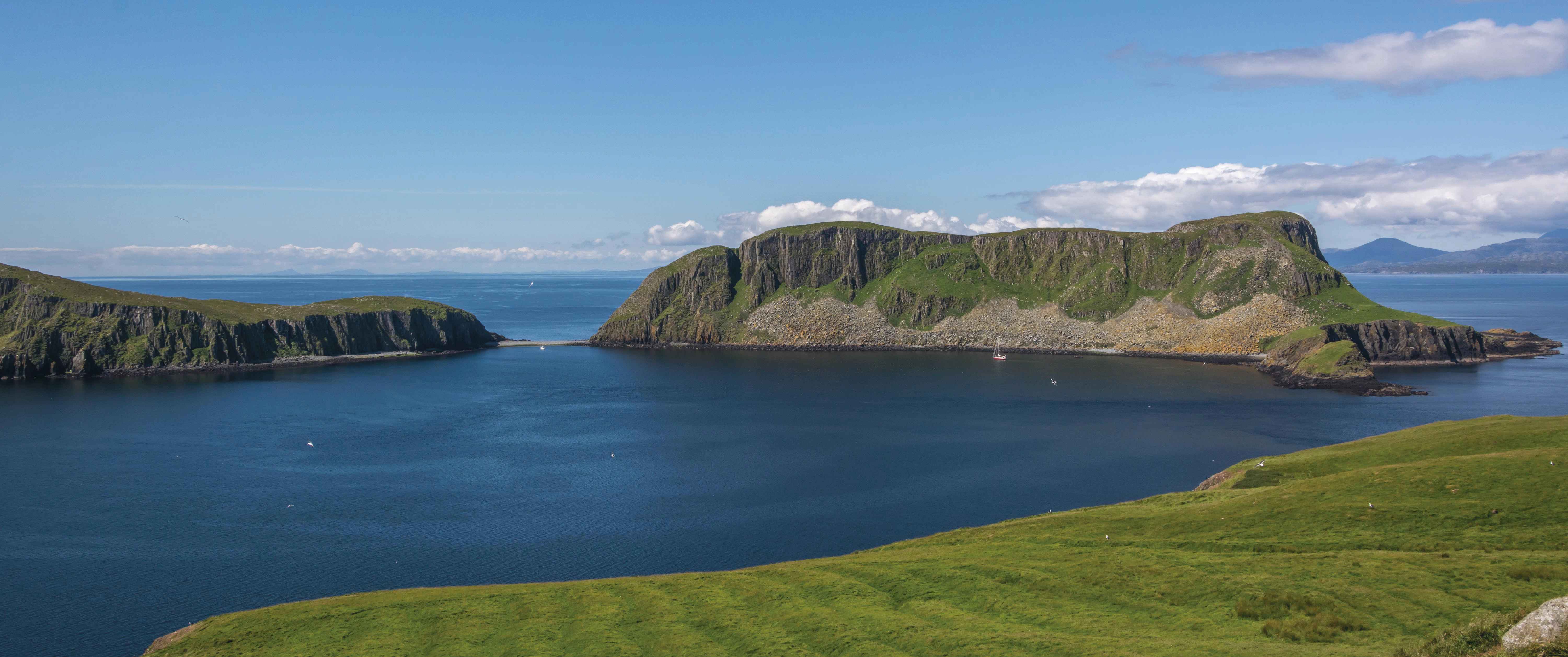 A series of islands, with steep cliffs, green grassy slopes and boulders sit surrounded by deep blue sea under a bright blue sky with a few white clouds. 